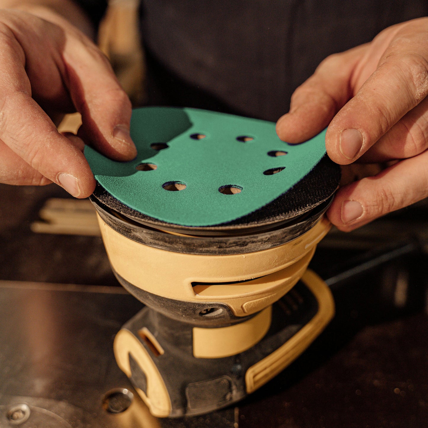 Woodworker fitting a green aluminum oxide 8-hole sanding disc onto a random orbit sander pad