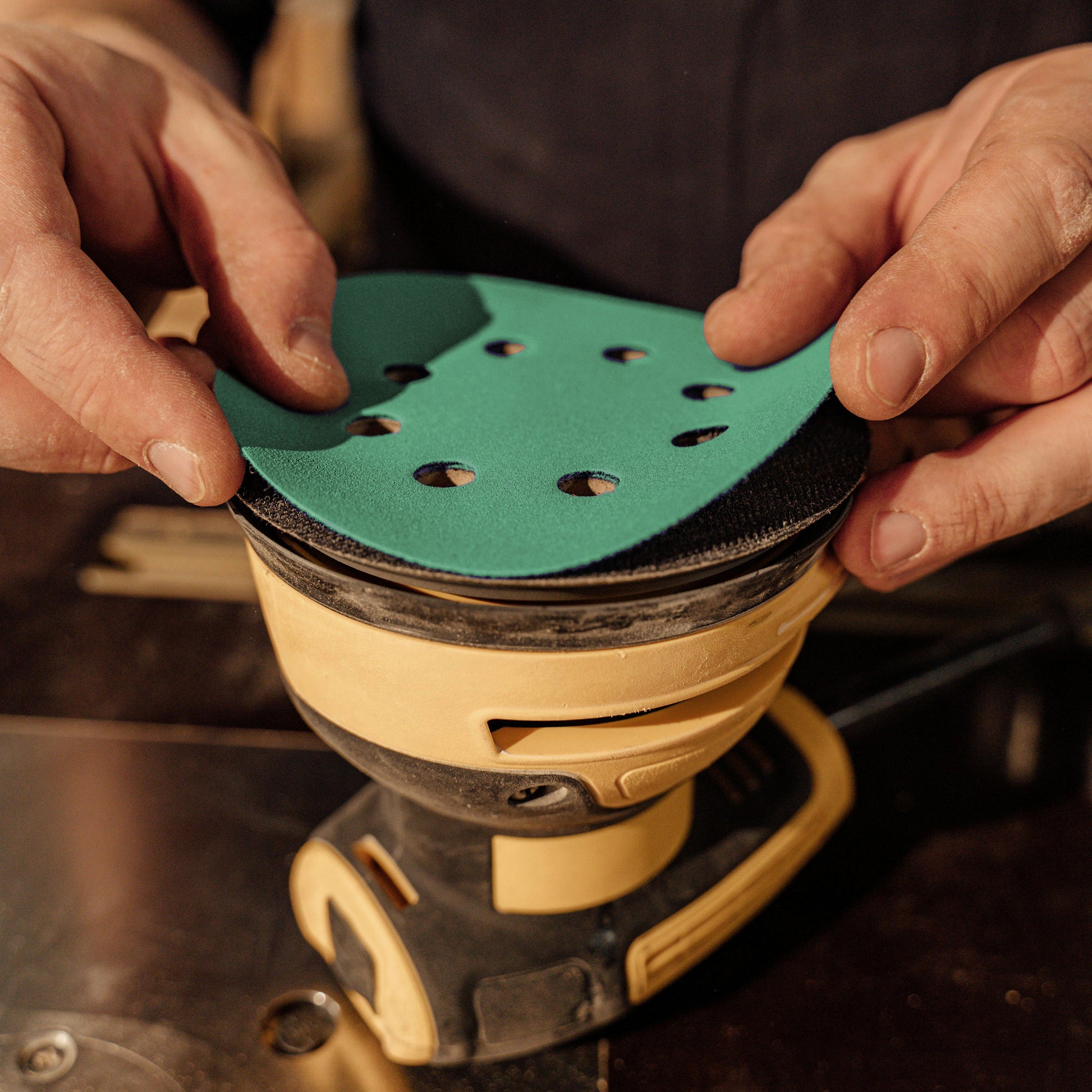 Woodworker fitting a green aluminum oxide 8-hole sanding disc onto a random orbit sander pad